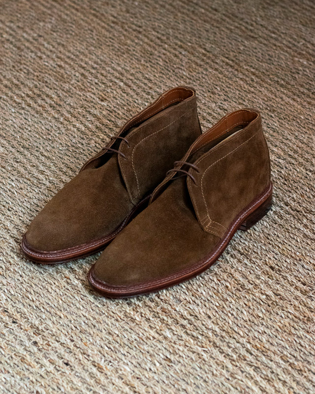 A pair of brown suede chukka boots with dark brown laces, resting on a textured beige woven rug.