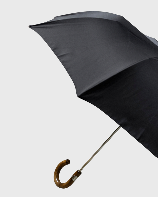A close-up of a black umbrella canopy with a curved brown wooden handle and silver metal shaft, shown against a plain light gray background.