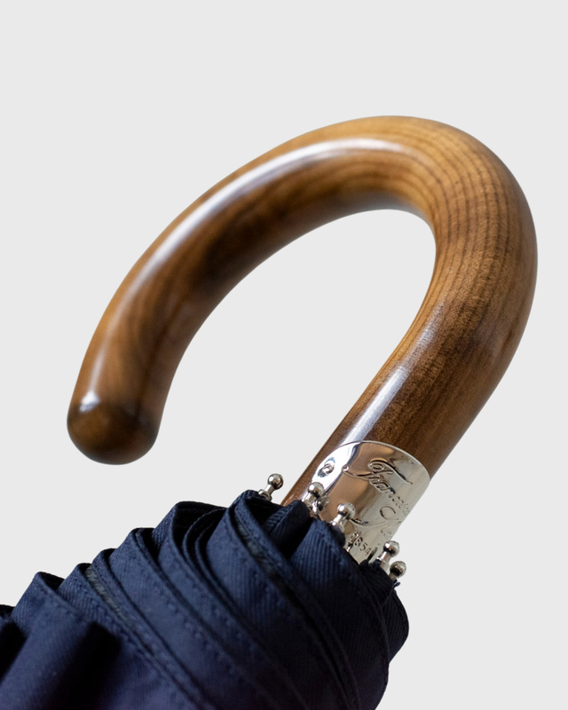 Close-up of a luxury umbrella handle made of polished brown wood, featuring a silver metal collar with decorative rivets and a navy blue fabric canopy, set against a light gray background.