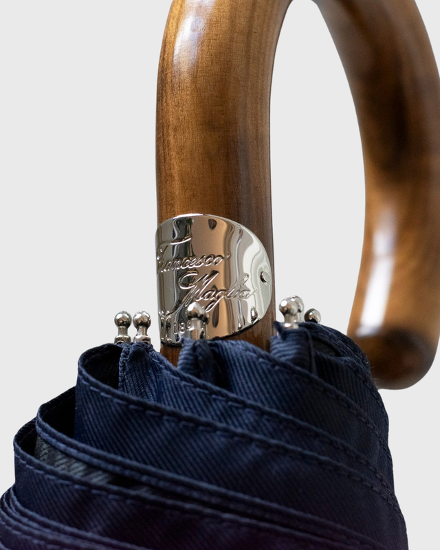 Close-up of a folded umbrella handle made of polished brown wood, secured with a silver metal band engraved with the brand name 'Tuscany Japan', and topped with navy blue fabric.